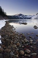 Rocks and snow of the shoreline of a placid lake with the sun just lighting up the distant mountains